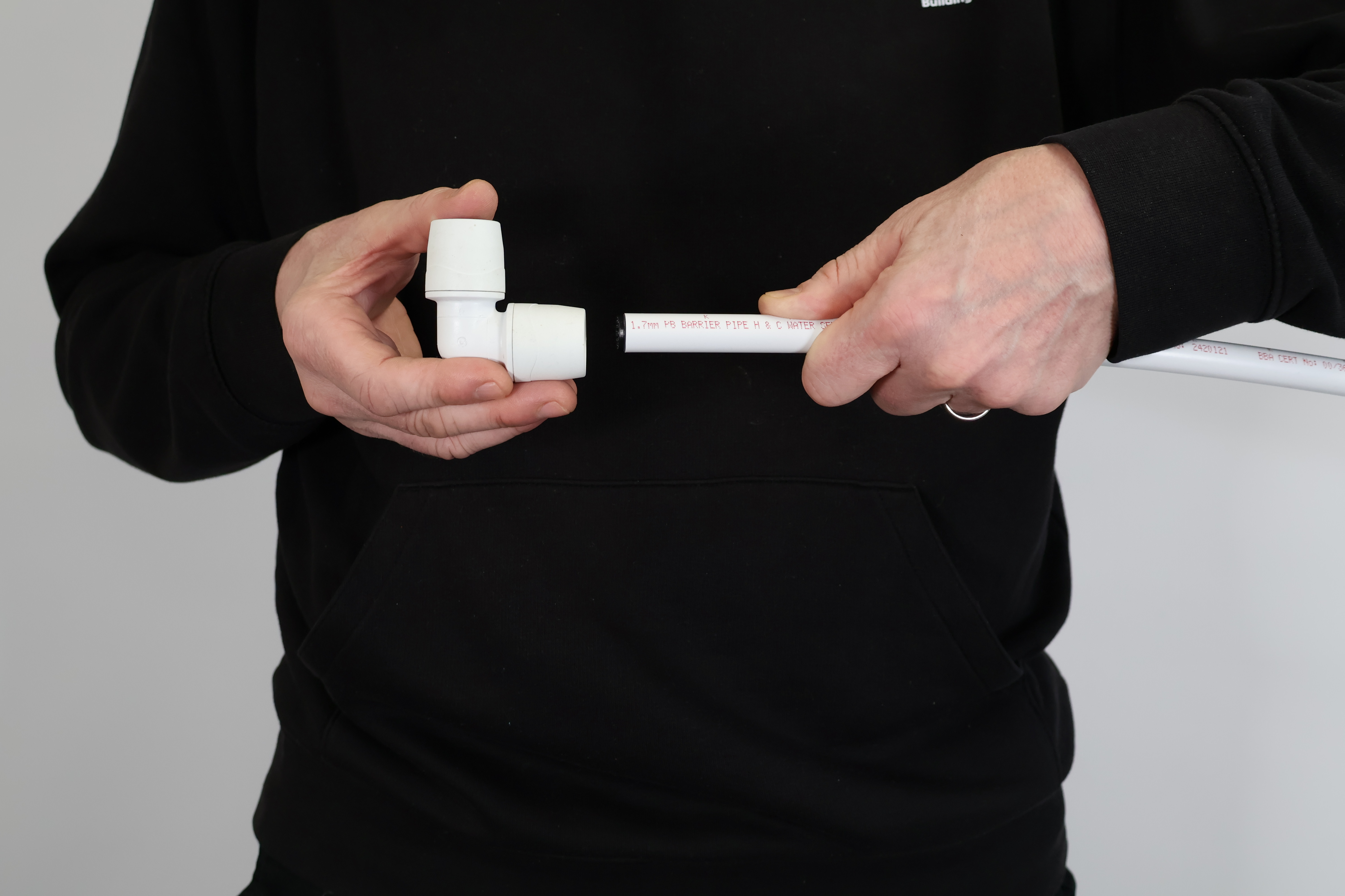 Close-up showing the insertion of a plastic plumbing pipe into a PolyMax push-fit elbow. The pipe is prepared with a square cut and internal stiffener to ensure it seats correctly against the internal O-ring for a watertight seal.