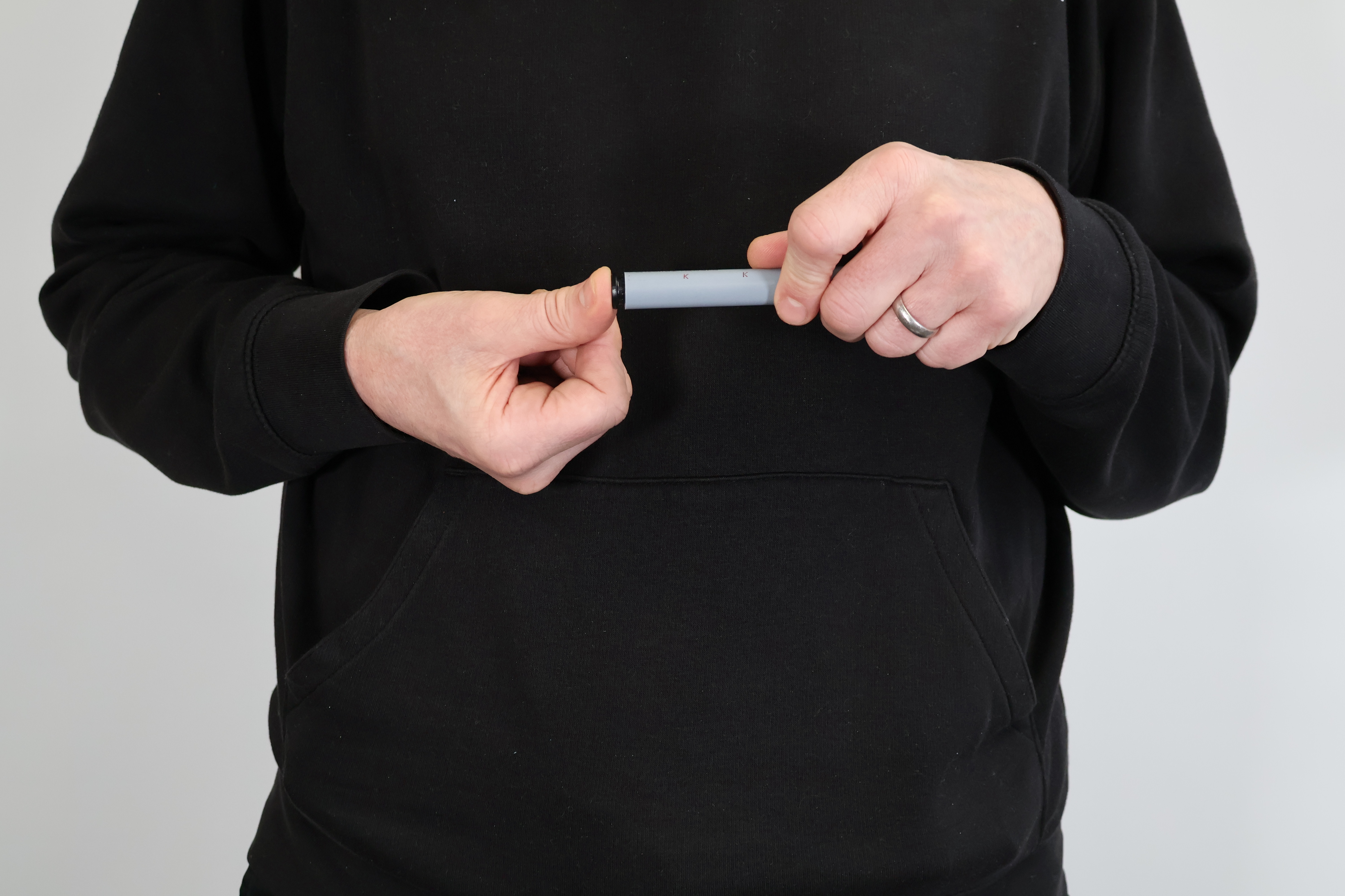 A medium close-up photograph against a light grey background, showing a person in a black hoodie and a wedding band, demonstrating the precise insertion of a small black plastic pipe stiffener (sleeve) into the end of a grey PolyPlumb polybutylene push-fit plumbing pipe. This image captures the critical preparation step for ensuring a secure and leak-free joint, suitable for use as a technical guide or product page element.
