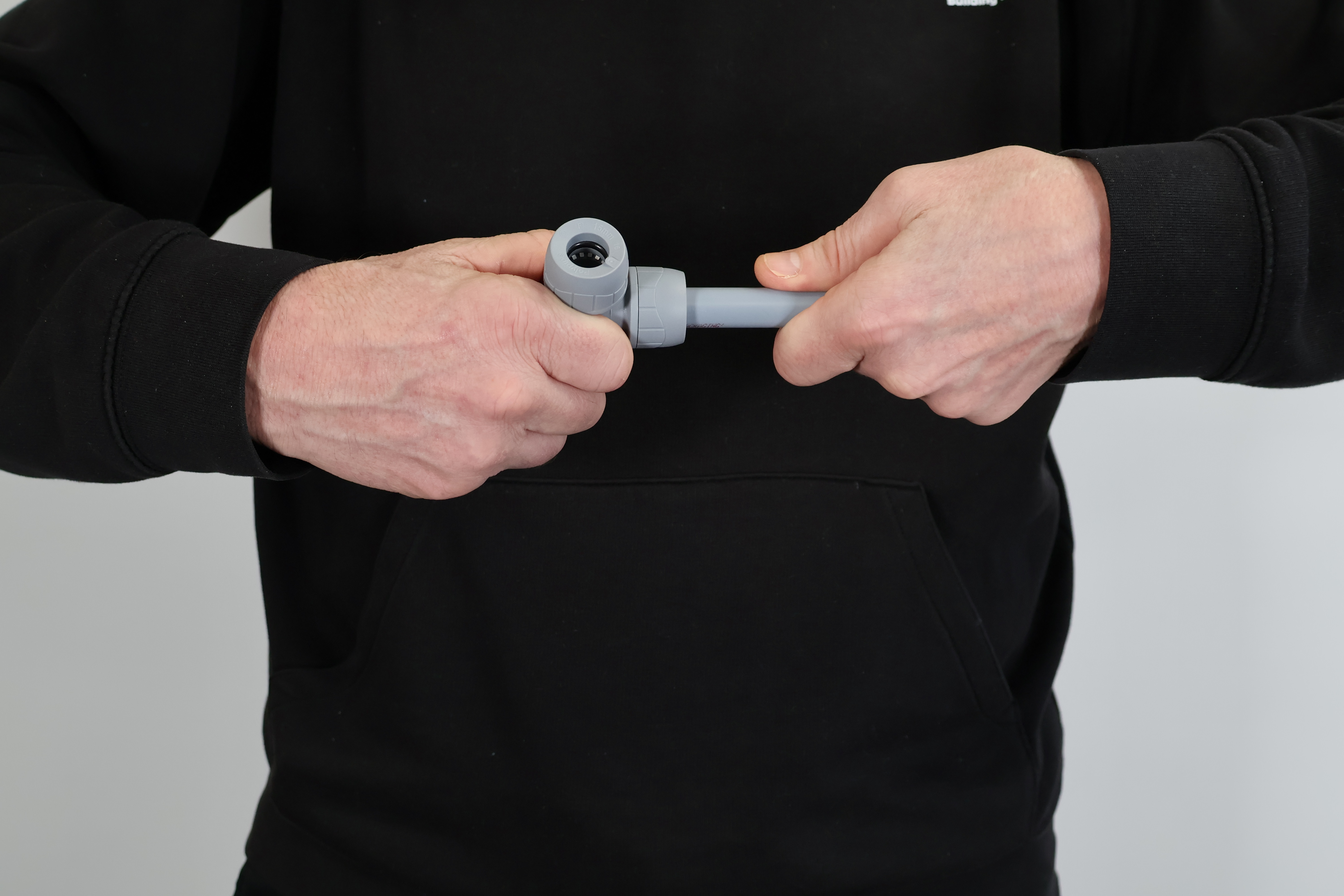 A close-up photograph of a person in a black hoodie completing the connection of a grey PolyPlumb push-fit elbow fitting to a length of polybutylene pipe. The image shows the final "push-home" action, illustrating a secure, tool-free plumbing installation. This professional demonstration highlights the ease of use and reliability of the PolyPlumb plumbing system for DIY or trade applications.