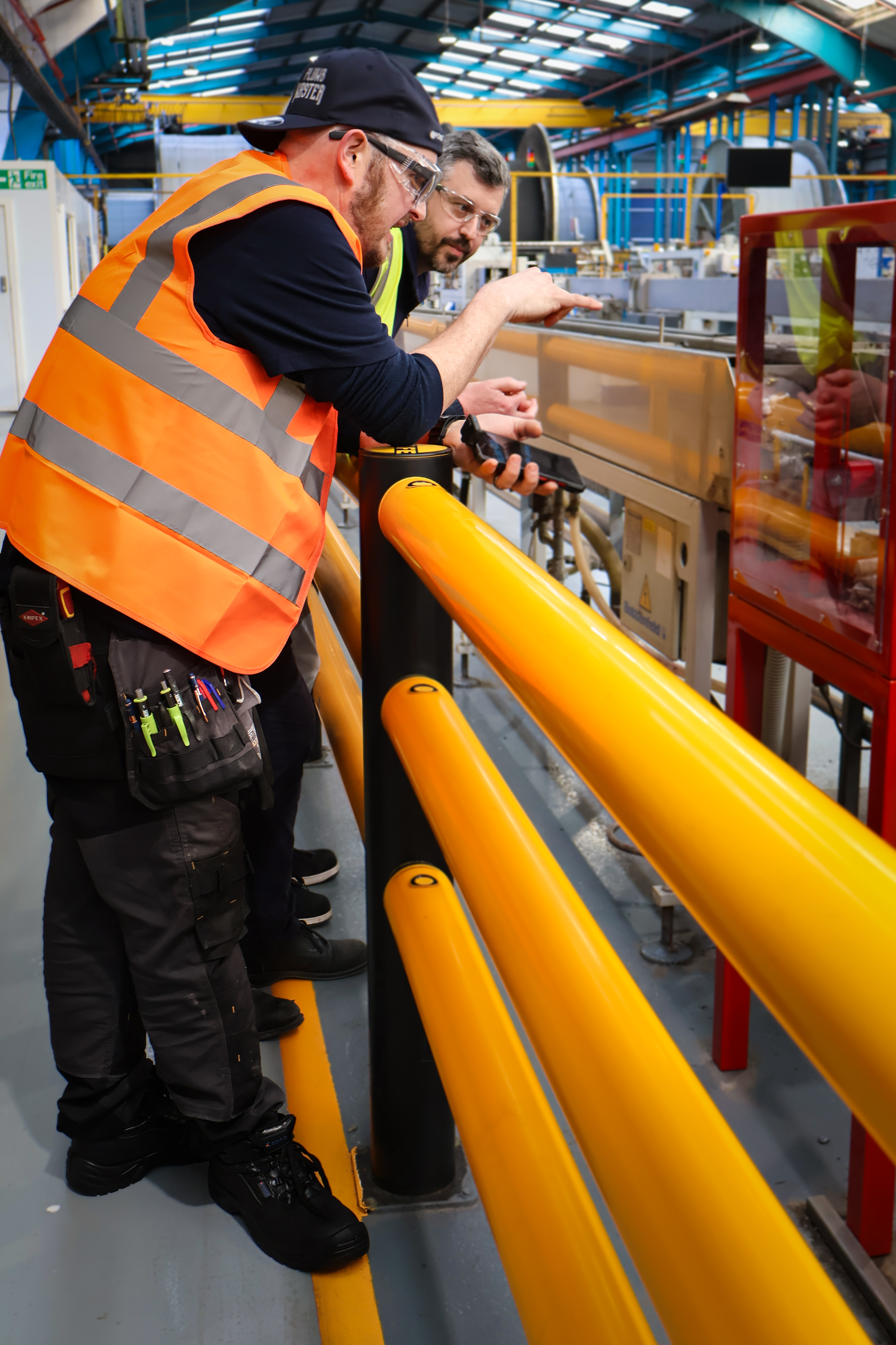 Inside the Polypipe Building Products manufacturing facility, Chris Rowland stands alongside Pointy Plumber to explain the operation of the high-speed pipe printing machine. Pointy, dressed in his signature orange high-visibility vest, dark work trousers with a loaded tool pouch, and a "Plumb Master" cap, leans over a bright yellow safety railing while holding his smartphone to record the process. Chris, also in safety gear, points toward the machinery where informational text is being printed onto white polybutylene pipe as it moves through the production line. The background reveals the vast scale of the factory, with overhead skylights, industrial racking, and large coils of piping stretching into the distance.