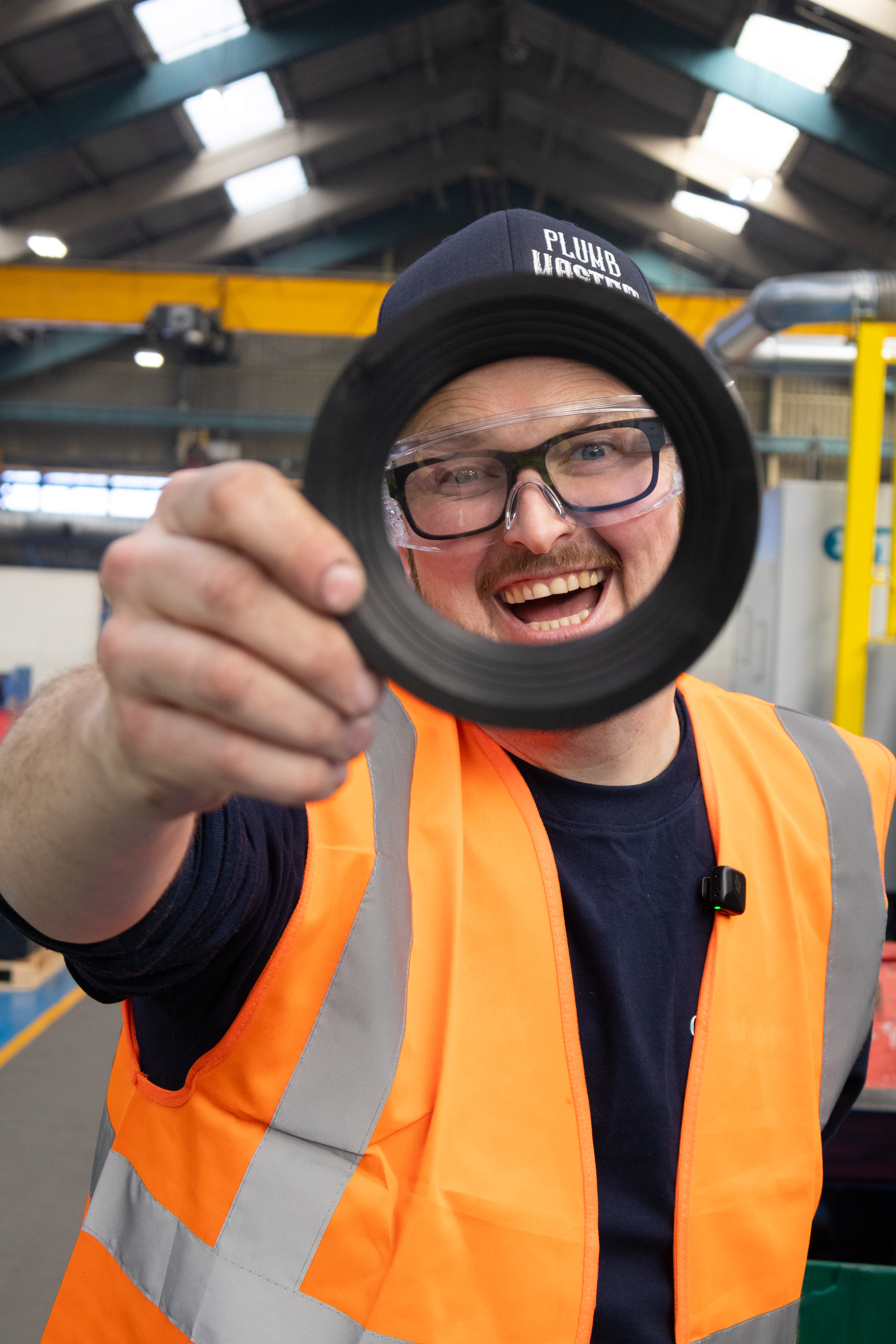 A close-up photograph of Pointy Plumber in a factory, playfully framing his smiling face by holding a large, black rubber seal from a pan connector directly toward the camera lens. He is wearing a navy blue shirt, a bright orange high-visibility vest, clear safety glasses, and a dark cap with "PLUMB MASTER" text. His expression is joyful and enthusiastic as he looks through the circular opening of the seal. The background shows the high-ceilinged industrial interior of the Polypipe Building Products factory, with large overhead beams, industrial lighting, and machinery visible in a soft focus.