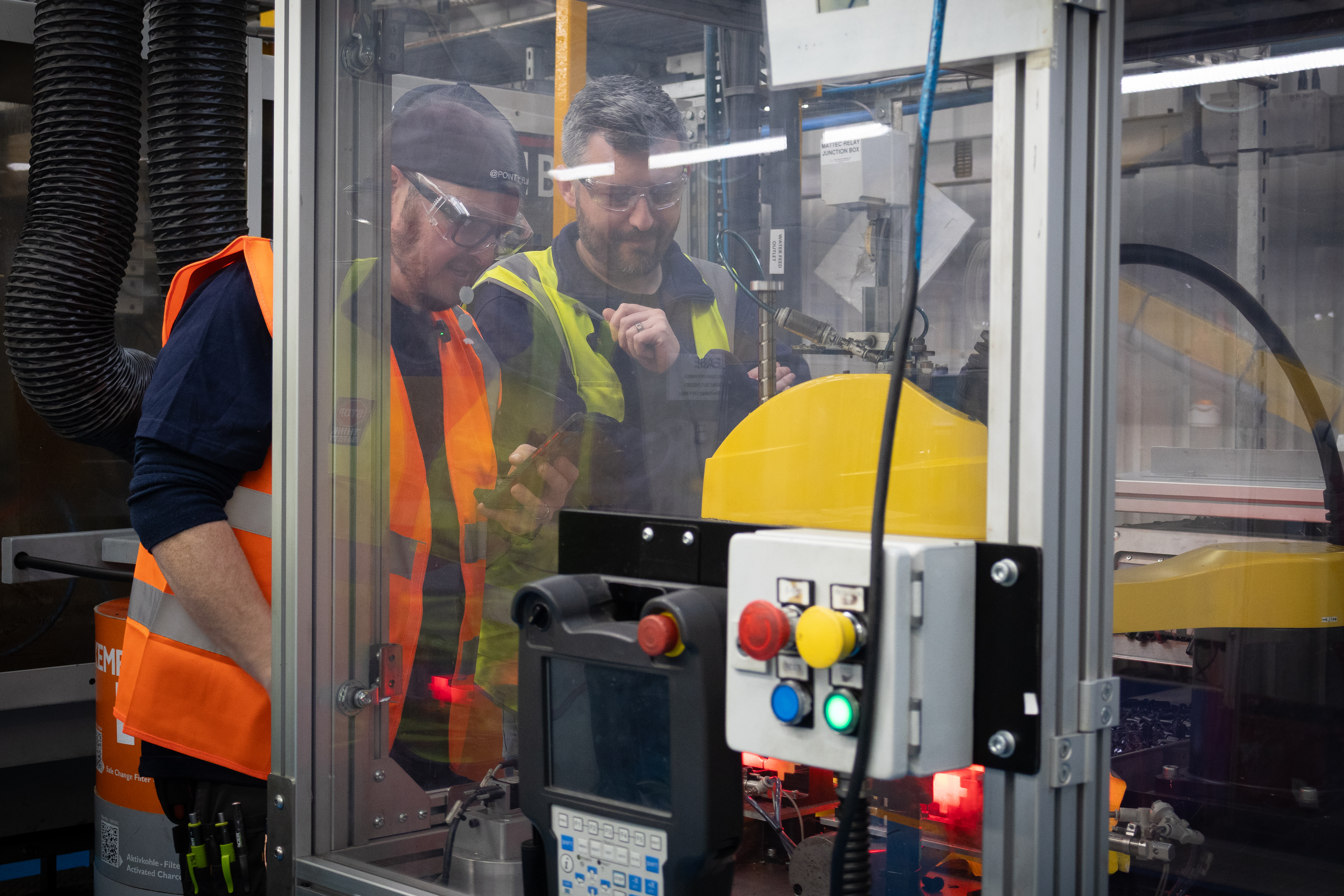 In a factory setting, Pointy Plumber, acting as the plastic plumbing ambassador for Polypipe Building Products, stands alongside a colleague as they observe a quality control machine through a clear safety glass panel. Pointy, wearing a navy blue long-sleeved shirt, an orange high-visibility vest, safety glasses, and a dark cap, holds a smartphone as he looks intently at the automated process inside. The machine is specifically designed to quality check In-Cert pipe stiffeners, featuring a yellow component and a control box with illuminated red, yellow, blue, and green buttons in the foreground. The industrial environment includes thick black ventilation ducting to the left and various technical components visible through the protective enclosure.
