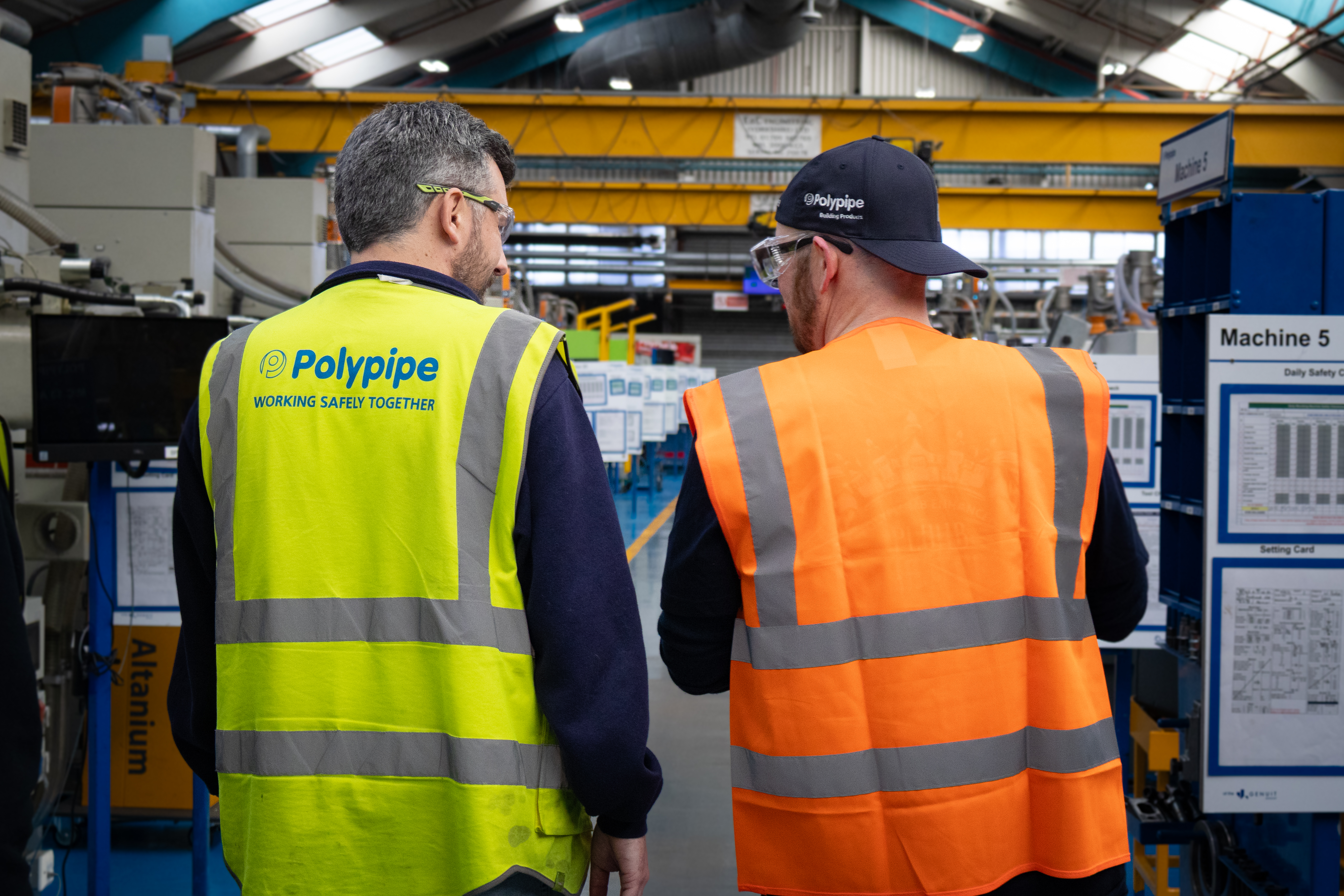 A medium shot from behind shows Pointy Plumber, plastic plumbing ambassador for Polypipe Building Products, walking through the large, industrial extrusion hall alongside Development Engineer Chris Rowland. Pointy is on the right, wearing a bright orange high-visibility vest and a dark blue cap with the Polypipe logo. Chris walks on the left, wearing a yellow high-visibility vest featuring the Polypipe logo and the slogan "Working Safely Together". They are surrounded by various manufacturing machines, including one labeled "Machine 5," with yellow gantry cranes and industrial ducting visible in the high-ceilinged background.