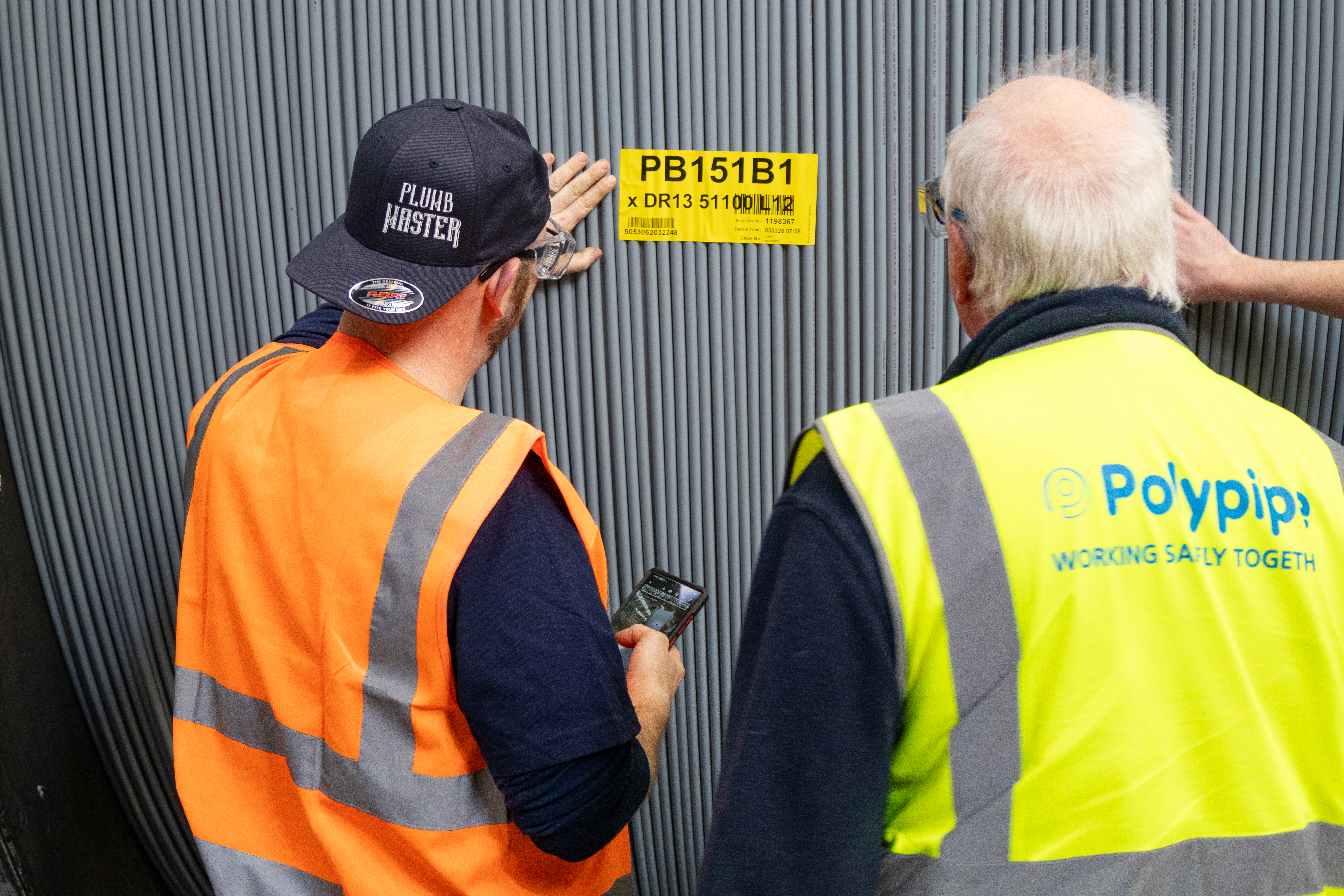 In a close-up photograph taken at the Polypipe Building Products factory, Pointy Plumber and Quality Assurance Engineer Marcus Bennett are seen from behind, inspecting a massive coil of grey plumbing and heating pipe. Pointy, wearing his signature orange high-visibility vest and a black "Plumb Master" cap, holds a smartphone in his right hand while reaching out with his left to touch the densely packed vertical rows of polybutylene pipe. Marcus, wearing a yellow high-visibility vest with the Polypipe logo and the slogan "Working Safely Together," stands to his right. A yellow identification label marked "PB151B1" is affixed to the side of the giant coil, which is currently undergoing a three-day curing process.