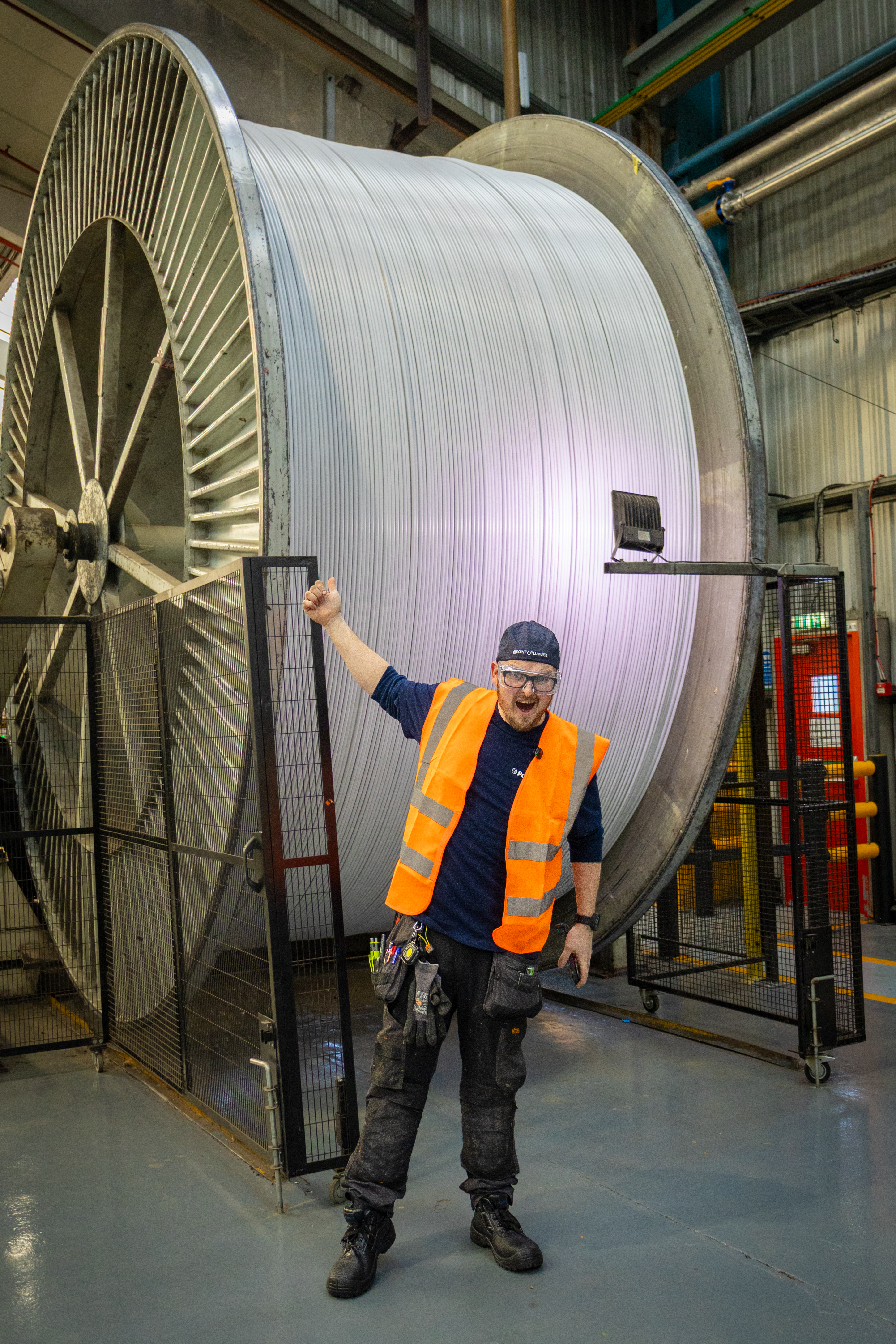A full-length photograph taken within an industrial factory shows Pointy Plumber, Polypipe Building Products plastic plumbing Ambassador, giving an enthusiastic thumbs-up in front of an immense, towering metal spool full of polybutylene pipe. The worker, dressed in a navy blue shirt, an orange high-visibility vest, dark trousers, a tool belt with a multi-tool, safety glasses, and a dark blue cap, is smiling broadly at the camera with his left arm raised. The gigantic spool behind him is spooled with bright, grey pipe, its scale making him look very small. 