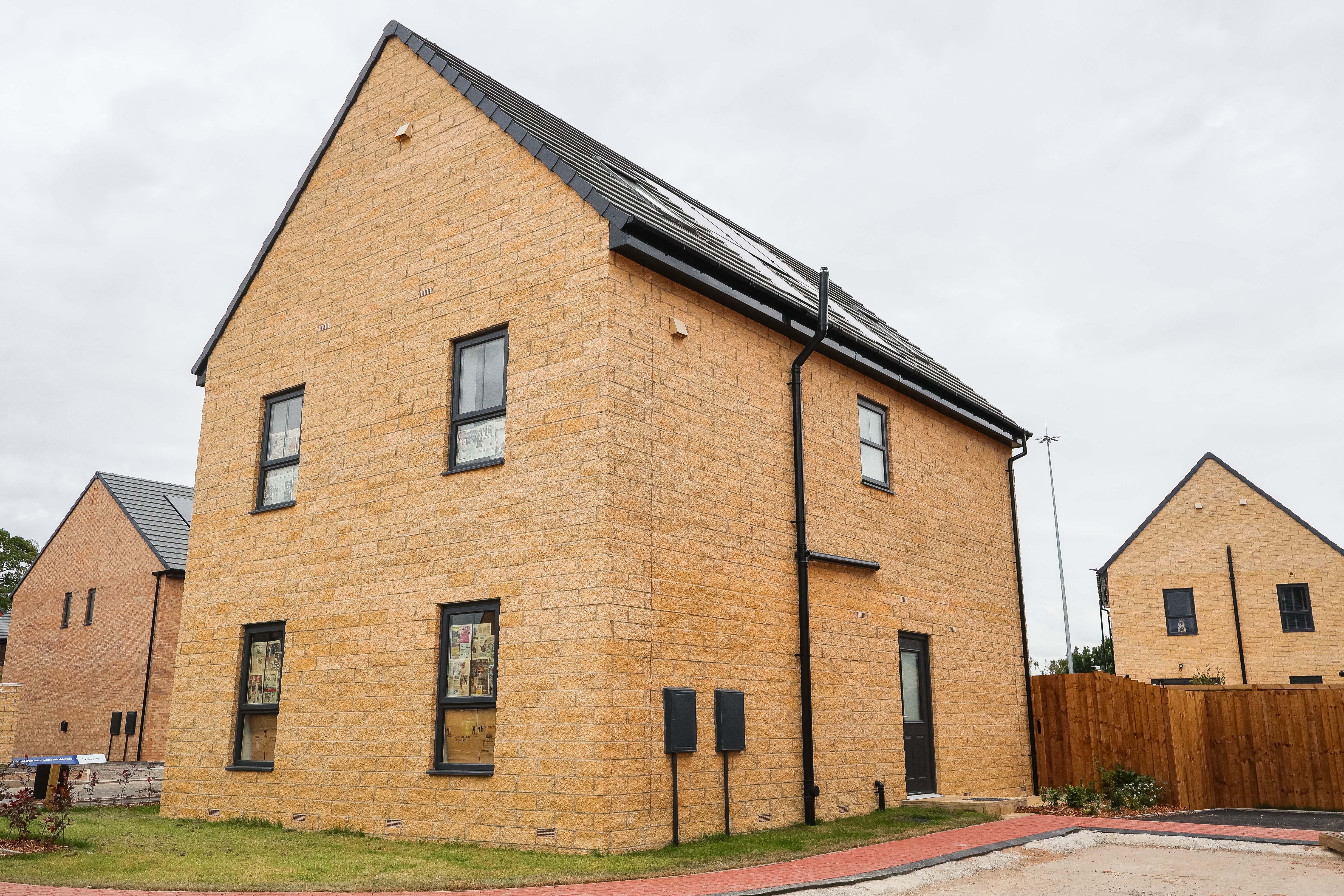 The image showcases a newly constructed home with a black vertical soil stack and matching guttering installed against light-colored textured brickwork. The house features anthracite grey window frames, a black side door, and solar panels integrated into a slate-grey tiled roof. This photo highlights contemporary residential architecture and the external application of Polypipe drainage solutions in a new-build housing development.